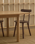 The Wooden Stories Itamae Dining Table in Light Oak is paired with two dark-seated wooden chairs on a woven rug, set against a slatted wall in a minimalist room, highlighting classic Danish furniture style.