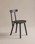 The Figura Dining Chair by Wooden Stories, shown in black oak with a rounded seat, three straight legs, and a curved backrest on a beige background, is ideal for contract-grade spaces and modern interiors.