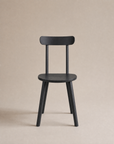 The Figura Dining Chair | Black Oak by Wooden Stories is a sleek, modern Scandinavian piece with four legs, a round seat, and a flat backrest—ideal for contract grade spaces and shown against a plain beige background.