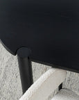 A close-up, top-down view of the Wooden Stories Cove Dining Table - Black Oak and a cream textured armchair on a striped light gray carpet creates an inviting dining scene with organic design.