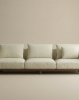 The Islet Sofa | Fabric by Wooden Stories is a modern beige sofa with three seat cushions, four back cushions including a smaller one, set against a plain, light-toned wall and floor background.