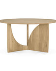 The Ethnicraft Geometric Dining Table - Oak is a round, solid oak table with a smooth top and distinctive base featuring intersecting curved and angled panels in a light natural wood finish.