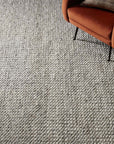 The Hendrick Rug - Grey by Loloi, a textured light gray hand-woven wool area rug with a pebble pattern and GoodWeave certified, is pictured with a rust-colored armchair and a small round side table in the corner.