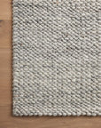 A close-up of the Loloi Hendrick Rug - Grey, a chunky hand-woven wool area rug with a textured, pebble-like pattern, covers most of a light brown wooden floor. One edge of the rug is visible.