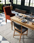A dining room features a wooden table set for four with plates, glasses, bread in a basket, and woven placemats. An orange armchair and a wooden chair sit on the Loloi Hendrick Rug - Grey over black-and-white tile flooring.
