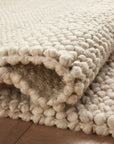 A close-up of the Hendrick Rug - Natural by Loloi, a chunky hand-woven wool area rug in beige, partially folded on a wooden floor to highlight its thick texture and soft woven pattern.