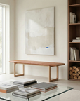 A modern, minimalist living room showcases the Hover Bench by Wooden Stories with a leather seat, a large neutral-toned abstract painting with blue accents, stacked books on a glass coffee table, built-in shelves, and abundant natural light.