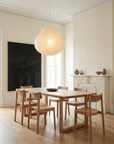 A minimalist dining room featuring the Wooden Stories Hover Dining Table in Light Oak with six matching chairs, a large paper lantern pendant, neutral walls, dark abstract art, and simple pottery on a white mantel.