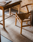 A wooden desk with an open magazine sits beside a woven chair on the Radhi Rug - Orchid by HIMALAYAN HERMITAGE, set against a rustic brick wall in a cozy, sunlit room.