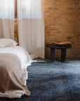 A cozy bedroom with a bed in beige and white linens, white curtains, a small wooden stool, brick walls lit by sunlight, and the Radhi Rug - Ink by HIMALAYAN HERMITAGE adding a blue hand-woven wool touch.