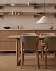 A minimalist dining room with Danish lines features the Hover Dining Table in Light Oak by Wooden Stories, four upholstered chairs, open shelves with ceramics, a pendant lamp, and neutral-toned furniture and decor.
