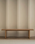 A minimalist room features the Wooden Stories Hover Bench | Leather Seat against a beige wall, with a dark wooden shelving unit displaying books and decor. A light rug on the wooden floor in the foreground adds warmth.