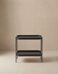 The Wooden Stories Humla Rectangular Side Table, a minimalist black piece with two shelves, features clean lines and modern Danish design inspired by Hans Bolling tray tables, shown against a plain beige background.