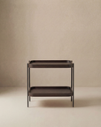 The Humla Rectangular Side Table by Wooden Stories features a minimalist two-tiered dark wood design with slim black legs and clean lines, inspired by Danish aesthetics. This modern piece stands elegantly against a beige background.