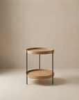 The Humla Round Side Table by Wooden Stories showcases Danish-inspired minimalism with a light wood top, lower shelf, and slender black metal legs, set against a neutral beige background.