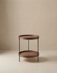 The Humla Round Side Table by Wooden Stories is a Danish-inspired wooden piece with two shelves and slim black metal legs, echoing Hans Bolling’s Tray Tables, shown on a plain beige background.