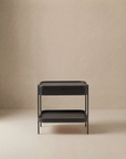 The Wooden Stories Humla Side Table With Storage features a minimalist black metal frame, solid wood drawer, and open lower shelf, shown against a plain beige background.