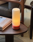 The Ami Table Lamp by Olive Studio x SOBU features a small, cylindrical red base and an origami-inspired pleated white shade, casting a warm glow on a dark wooden table beside a book and cushioned chair.