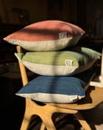 The Alma de Oaxaca Cushion - Sunset by SOBU, handwoven in Oaxaca using traditional techniques, sits on a wooden chair. Sunlight highlights its rich texture and colors, creating a cozy, inviting atmosphere.