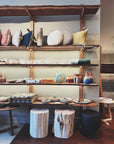 Wooden shelves showcase home decor—colorful cushions, mugs, ceramics, plates, and bowls. In front are two rustic stools and a black ottoman. SOBU Gift Cards by SOBU are available on the shelves for easy gifting.