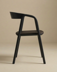 The Inlay Dining Chair by Wooden Stories features a minimalist profile in black oak and leather, with a curved backrest, photographed in profile against a beige background.