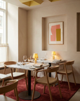 A modern restaurant interior features Wooden Stories’ Inlay Dining Chair in light oak and leather, marble tables with glassware and plates, small gold lamps, abstract wall art, a red rug, and sunlight streaming through the window.