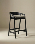 The Inlay Counter Stool by Wooden Stories features a modern black solid oak frame, curved backrest, footrest, and a dark leather seat, set against a neutral beige background.