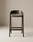 The Inlay Counter Stool by Wooden Stories features a solid oak frame, curved backrest, and round leather seat in dark brown, shown against a neutral beige background.