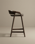 The Inlay Counter Stool | Leather Seat by Wooden Stories is a modern solid oak bar stool with a curved backrest and round leather seat, featuring smooth lines and minimalist design. Shown in profile against a beige background.