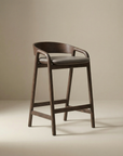 The Inlay Counter Stool by Wooden Stories features solid oak craftsmanship, a curved backrest, open sides, and a leather seat—an ideal modern addition for kitchen island seating against a plain beige background.