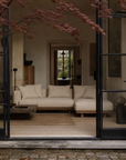 A serene, modern living room with the Islet Modular Sofa | Fabric • M1 by Wooden Stories in beige, durable upholstery, a wooden coffee table, and textured rug. Red-leaved tree frames open glass doors as natural light fills the space.