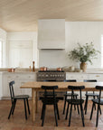 A modern kitchen with white cabinets, stainless steel stove, copper cookware, and the Wooden Stories Itamae Dining Table in light oak surrounded by six black chairs. A vase with green branches and earth-toned tiles complete the Scandinavian look.
