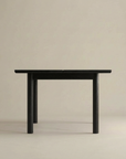 The Wooden Stories Itamae Dining Table | Black Oak features a minimalist Scandinavian-inspired design with four thick cylindrical legs, showcased against a plain beige background.