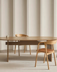 A modern dining room with a wooden oval table and four Wooden Stories Inlay Dining Chairs in light oak and leather, featuring plywood backrests and upholstered seats, a single cup on the table, light beige vertically lined walls, and a carpeted floor.