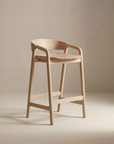 The Inlay Counter Stool | Wood Seat by Wooden Stories features a modern Danish design with a curved backrest, armrests, and footrest in smooth light wood—ideal for stylish kitchen island seating against a neutral beige background.
