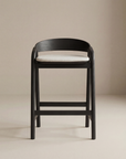 The Inlay Counter Stool | Fabric Seat by Wooden Stories features a modern black wooden frame, curved backrest, and light fabric cushion, shown against a neutral beige background.