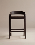 The Inlay Counter Stool | Wood Seat by Wooden Stories features modern Danish design in dark wood with a curved backrest and footrest, ideal for stylish kitchen island seating, shown here on a plain beige background.