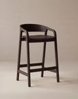 The Inlay Counter Stool | Wood Seat by Wooden Stories is a modern Danish counter stool in solid oak with a curved backrest and armrests. Ideal for kitchen island seating, this dark wooden piece has four legs with horizontal supports, shown on a beige background.