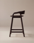 The Inlay Counter Stool | Wood Seat by Wooden Stories, featuring a modern dark wood design with a curved seat and backrest, is shown sideways against a beige background—ideal for stylish kitchen island seating.
