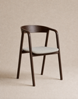 The Inlay Dining Chair | Fabric from Wooden Stories features a contemporary solid oak frame with a curved backrest and armrests, complemented by a light beige upholstered seat cushion. Inspired by the Danish Collection, this modern chair is set against a simple, solid beige background.