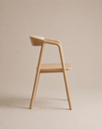 A minimalist wooden chair with smooth curves is shown in profile against a neutral beige background. The Inlay Dining Chair | Wood from Wooden Stories features a light wood finish, seamlessly blending form and function to suit contemporary interiors.
