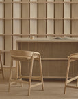 Three Wooden Stories Inlay Counter Stools with leather seats surround a minimalist solid oak counter topped with an open book and cup. An empty wooden bookshelf stands against a beige wall, softly lit in neutral tones.
