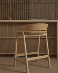 The Inlay Counter Stool by Wooden Stories, featuring a leather seat, is placed by a wooden desk with an open book and bowl, set against wood paneling and shelves in a warm, neutral-toned room.
