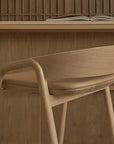 The Inlay Counter Stool with leather seat by Wooden Stories stands before a wooden desk. An open book and brown bowl are on the desk, while a wood panel wall forms a warm, inviting backdrop.