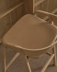 A modern Wooden Stories Inlay Counter Stool with a leather seat sits on a brown carpet, next to a matching solid oak desk.