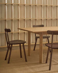Three wooden chairs surround the Wooden Stories Itamae Dining Table in Light Oak, set in a minimalist room with light wood panel walls and a beige woven rug, reflecting modern Danish design aesthetics.
