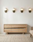 A minimalist living room with the SOBU Jupiter Sideboard - White Ash 3-door, a beige sofa, textured rug, and four modern wall sconces above the sideboard on a white wall.