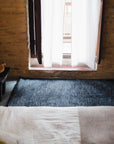 A softly lit room features a bed in the foreground, the HIMALAYAN HERMITAGE Radhi Rug - Ink on the floor, and sheer white curtains partially covering a window set in a rustic brick wall.