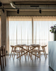 A modern dining area showcases the Ethnicraft Mikado Dining Table Round in oak, paired with wooden chairs. Sunlight streams through large windows with sheer curtains, while a vase of flowers adorns a nearby counter.