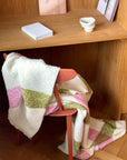 A pink bouclé chair is draped with the Elvang Denmark Lava Throw - Pink, a white Scandinavian blanket with green and pink stripes, in front of a wooden desk holding notebooks, a ceramic bowl, and photos propped at the back.